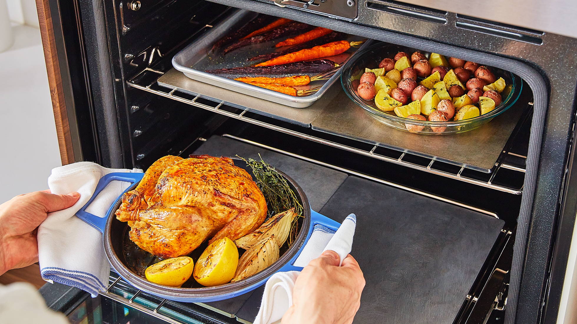 Hands place a roast chicken on a low oven rack atop a Misen Oven Steel. Two more Oven Steels are visible on the top rack beneath pans of vegetables.