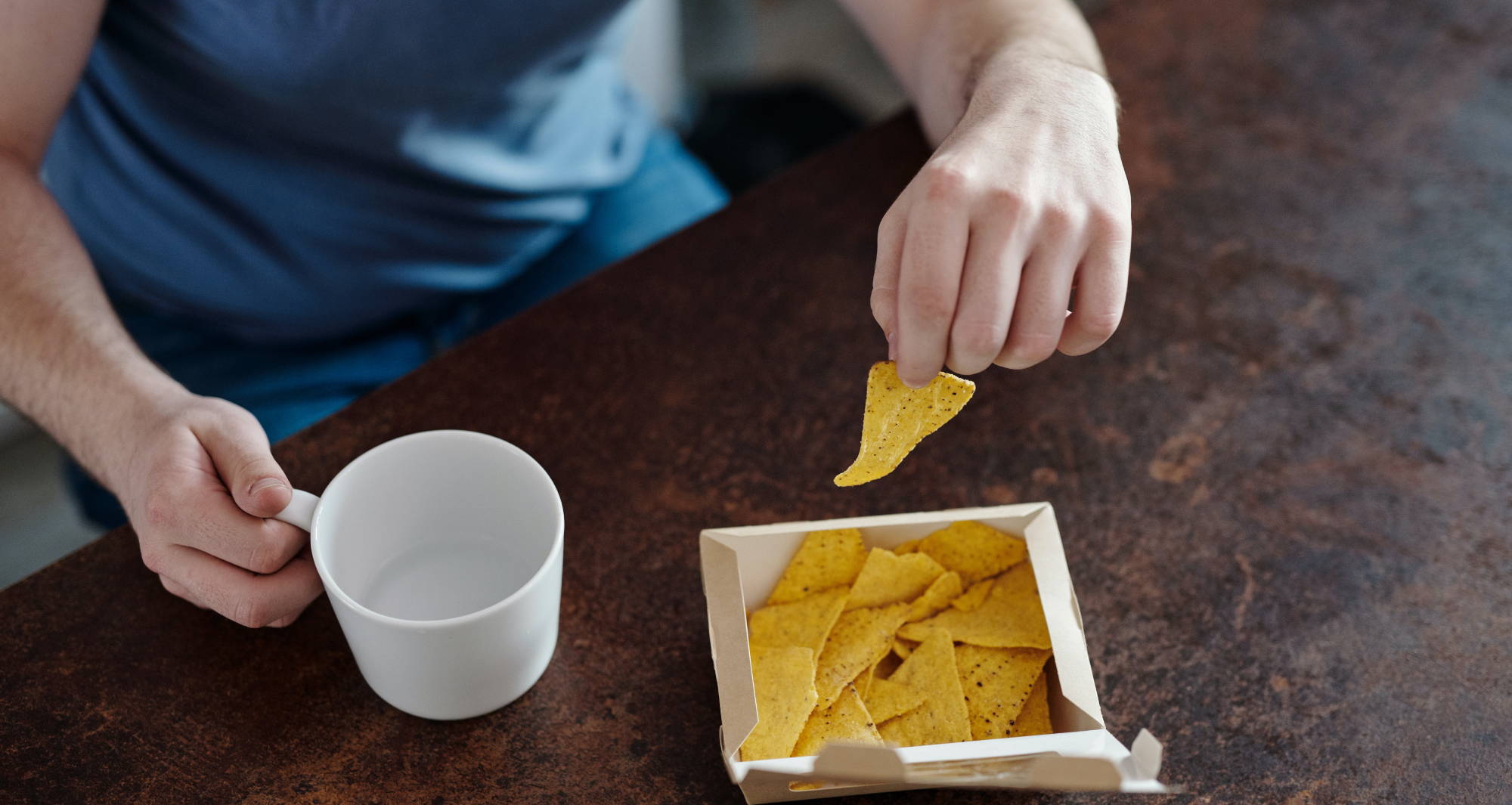 A man wearing a blue shirt holds a white mug and takes a tortilla chip out of a white paper box.