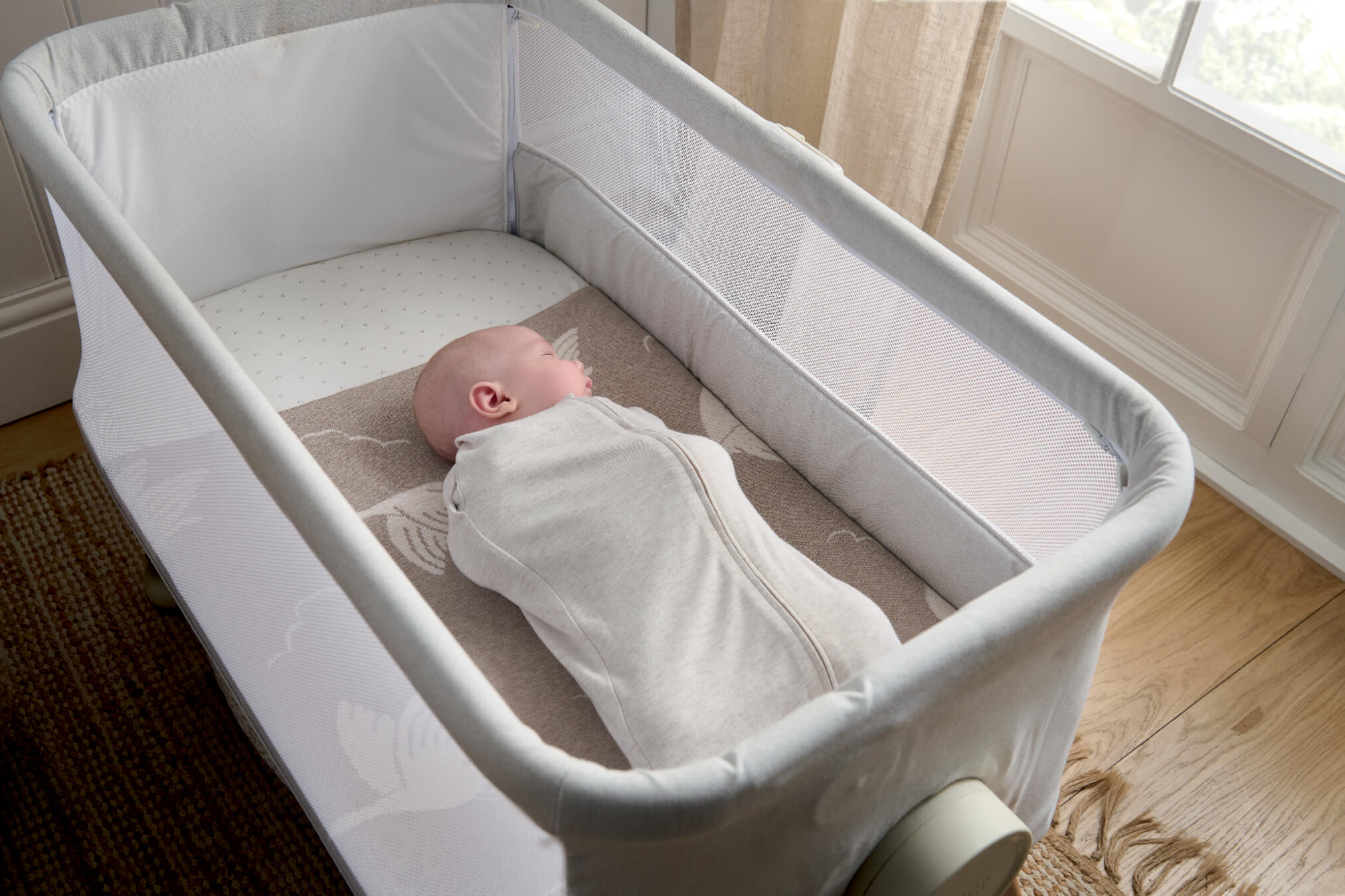 A newborn baby in an Oatmeal Swaddle in a bassinet near a window.