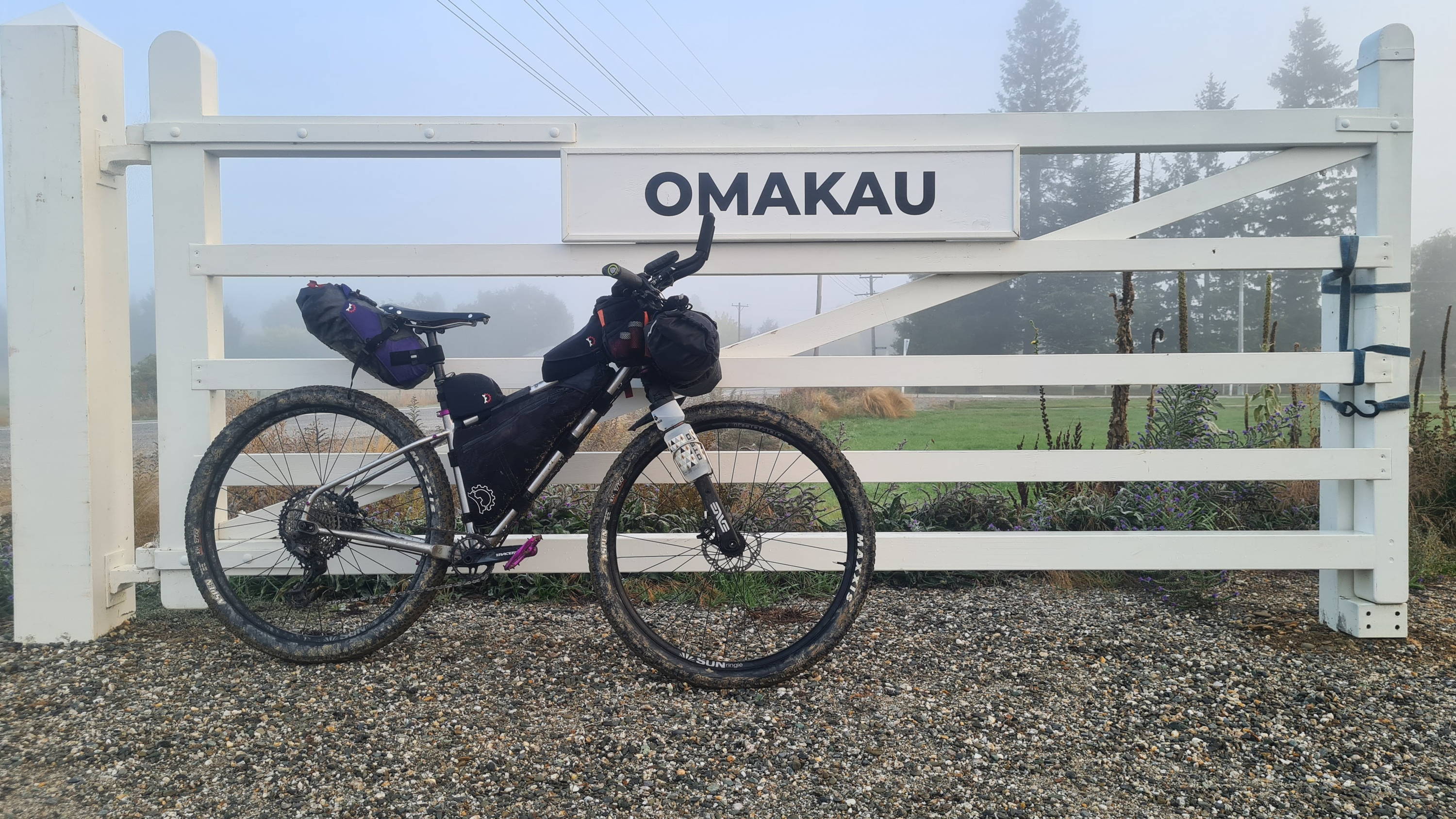 A fully-loaded Otso bikepacking bike leans against a white gate that reads 