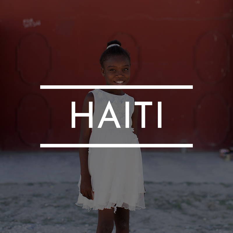 “HAITI” is written on top of an image of a young Haitian girl wearing white standing against a red gate