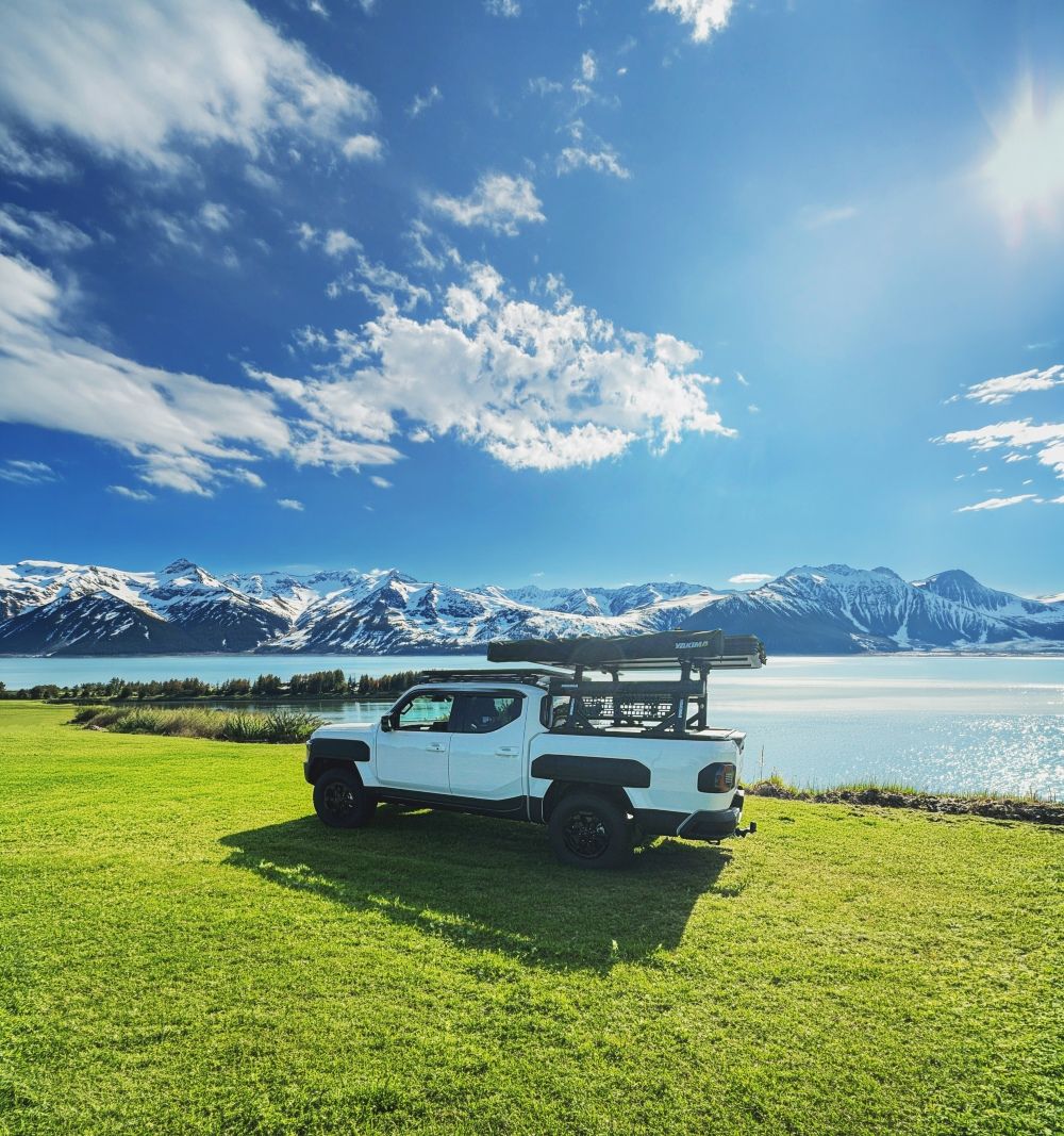 A Kia Tasman with Yakima Accessories, on green grass, in front of a lake and snow capped mountains