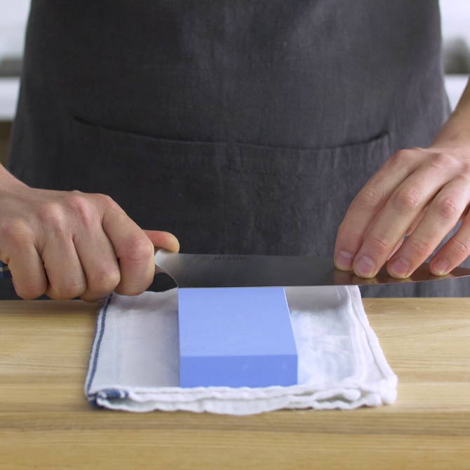 A chef sharpening a Misen Chef’s Knife on a blue 1000-grit Misen Sharpening Stone placed on a kitchen towel.