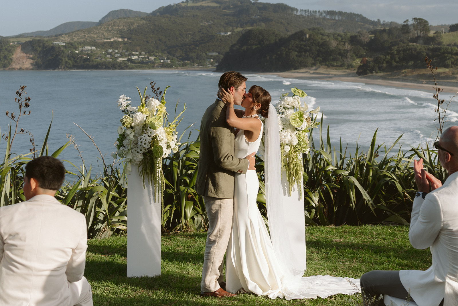 Bride and groom sharing a kiss at an outdoor wedding ceremony by the water.