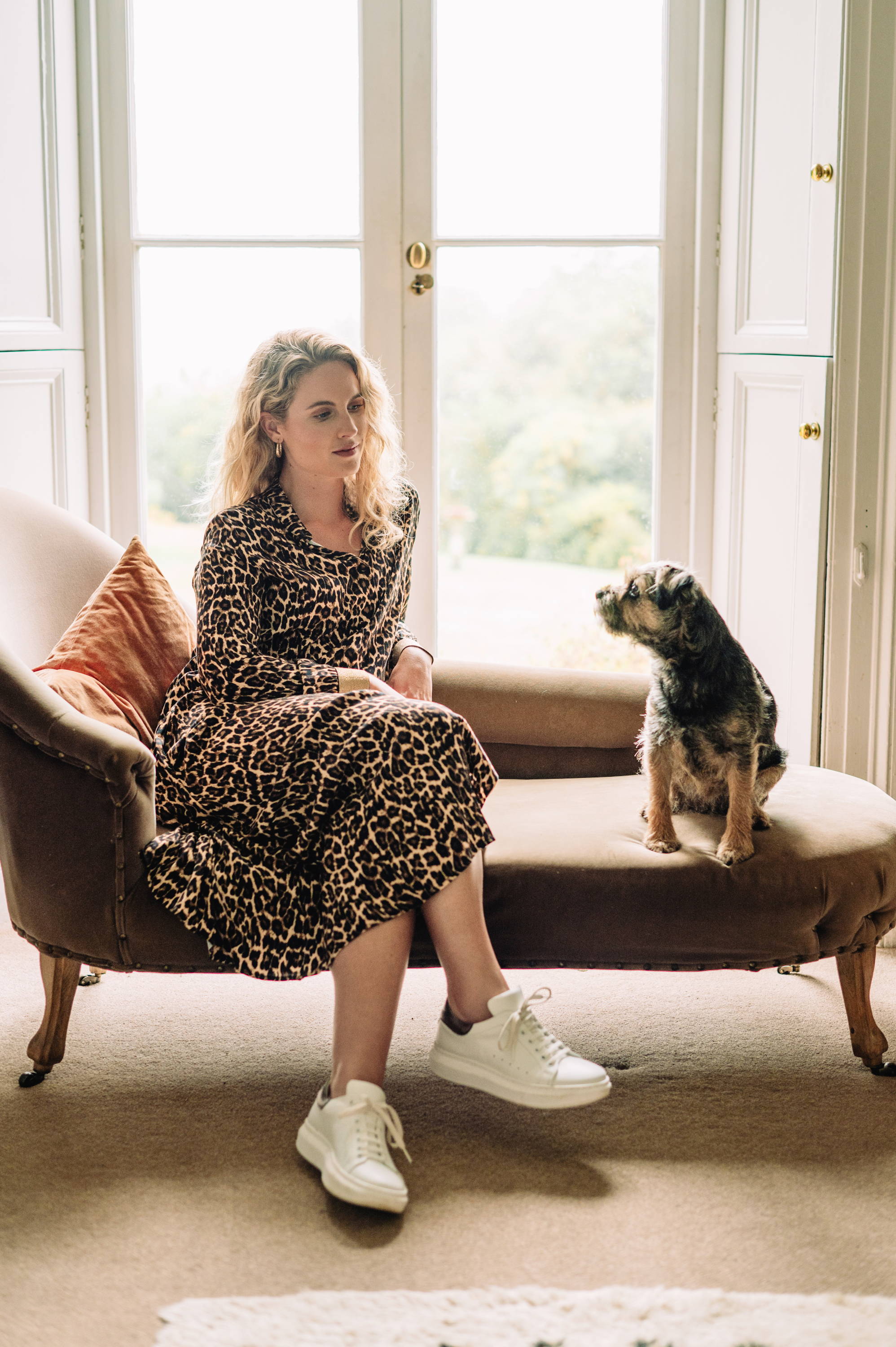 A model wearing a midi length leopard print dress with long sleeves and white trainers sitting on a golden chaise long with a small dog