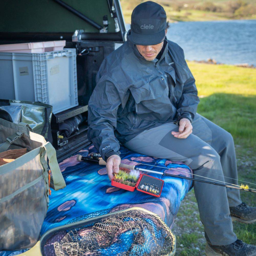 Chris Burkard wearing a grey rain jacket and grey pants sitting by the open trunk of a vehicle, with fishing gear spread out on the Thjorsa Original Puffy Blanket. 