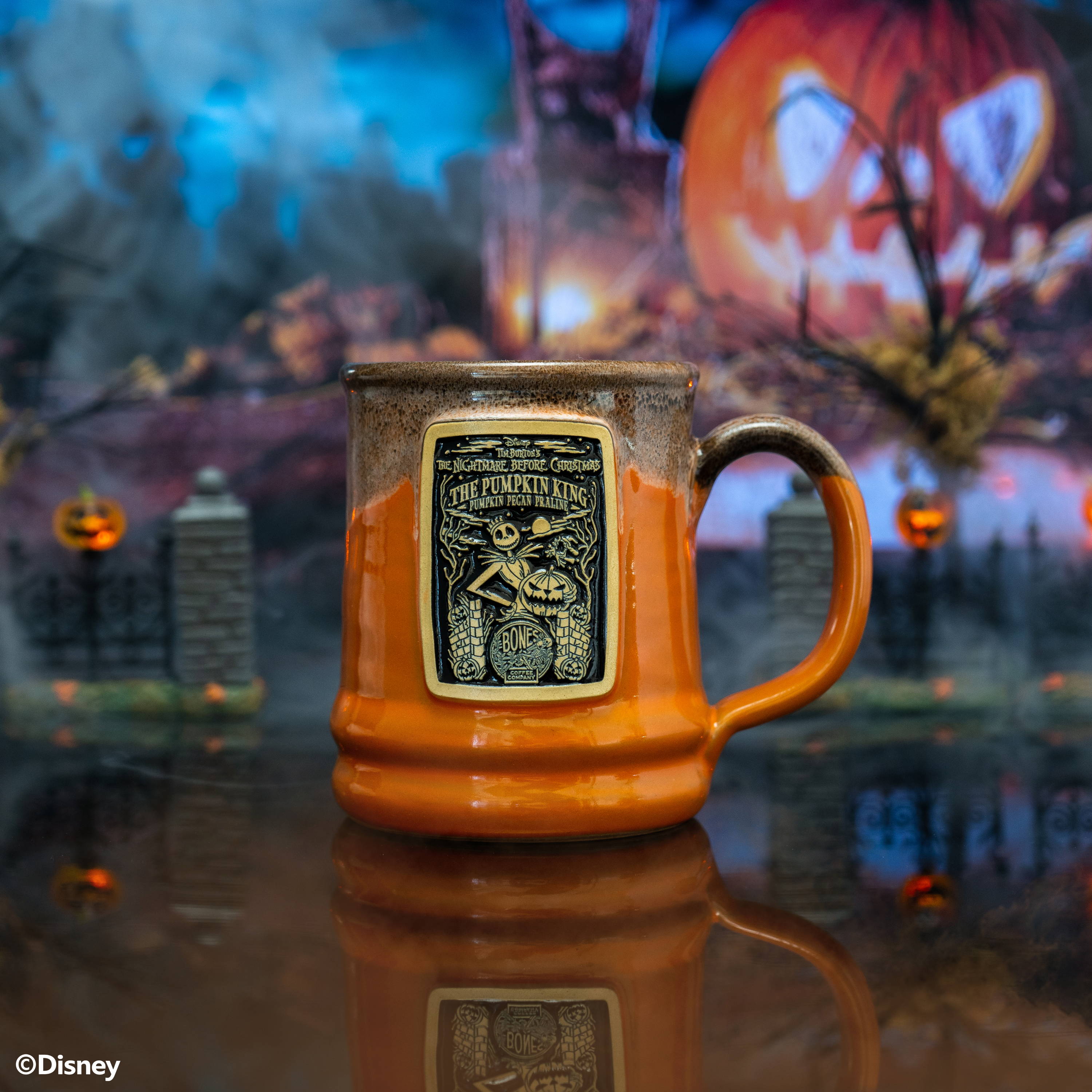 The front of the Bones Coffee Company The Pumpkin King hand thrown mug with Jack Skellington on the golden medallion. The mug is orange colored with a black and white glaze on top. It is inside a toy graveyard.