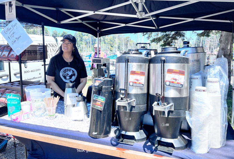 Jennifer Brown  behind the Thanksgiving Coffee Booth under a canopy with large coffee pots.