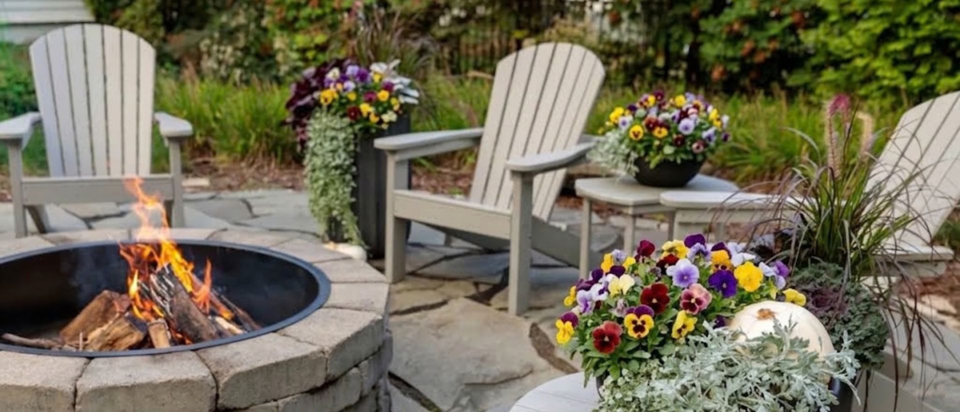 Scene of fall patio seating area featuring Adirondack chairs around a stone fire pit, surrounded by colorful pansies, ornamental grasses, and seasonal container plantings.