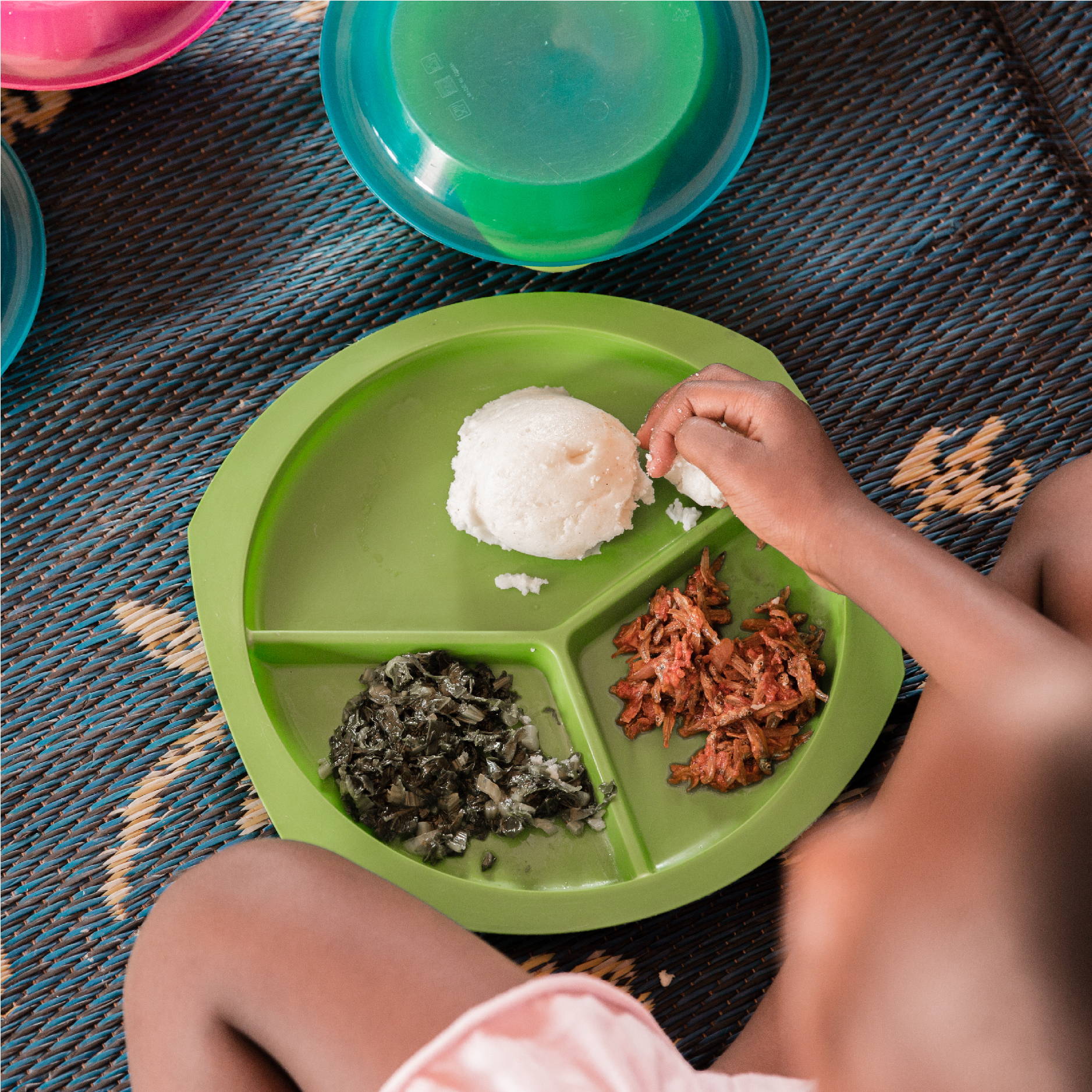 A girl is seen eating on a blue straw mat from a green plate. It is a traditional Zambian meal containing nshima 