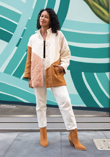 Woman with curly hair wearing a tan and brown jacket with cream colored pants and tan booties