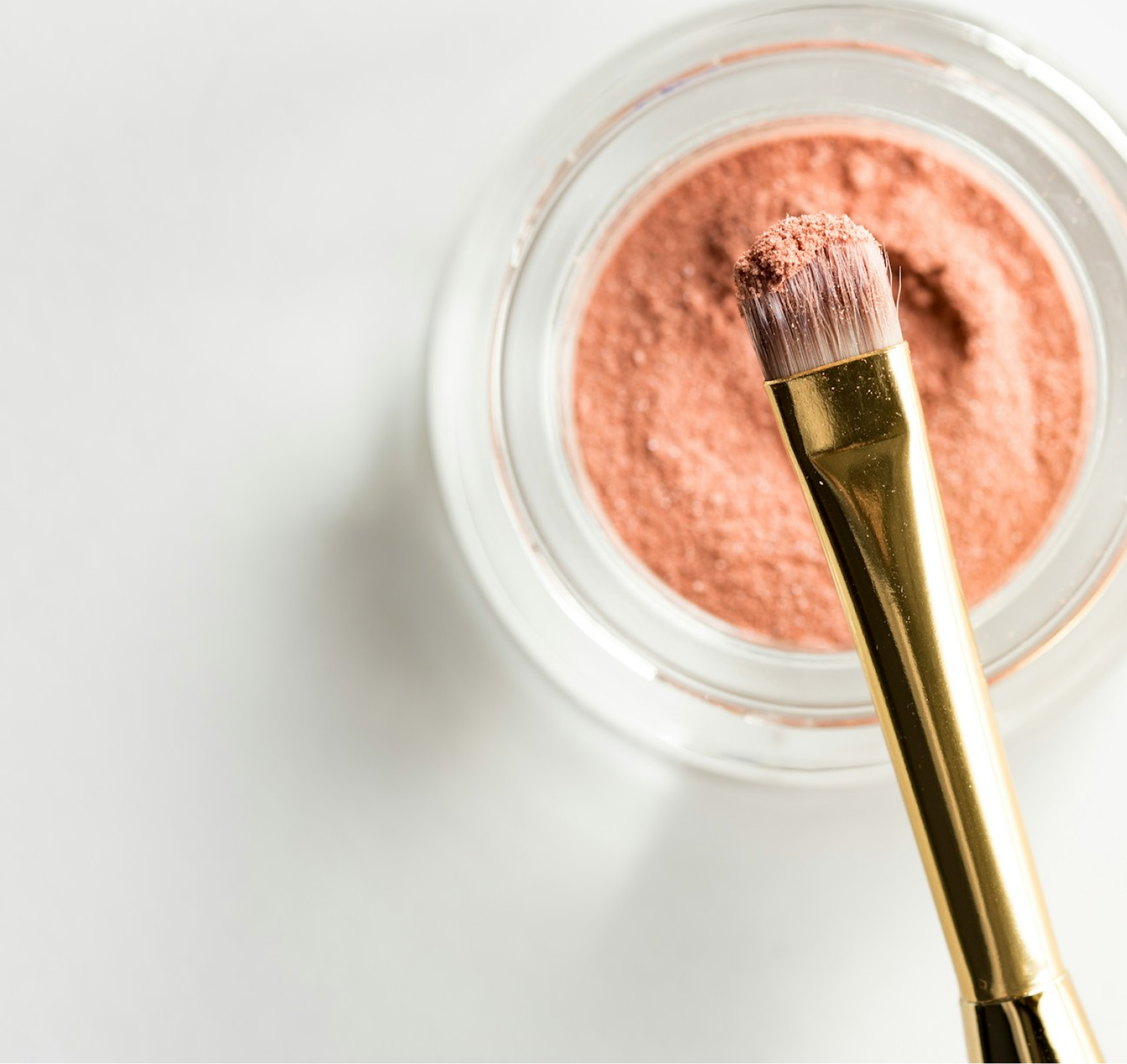 A glass pot containing makeup powder, with a gold-clad brush hovering over it, viewed top down.