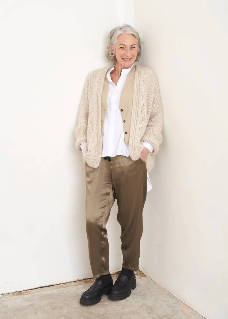 A model wearing a pair of khaki green satin trousers with a white top, khaki stripe waistcoat and an off white knitted oversized cardigan with black loafers