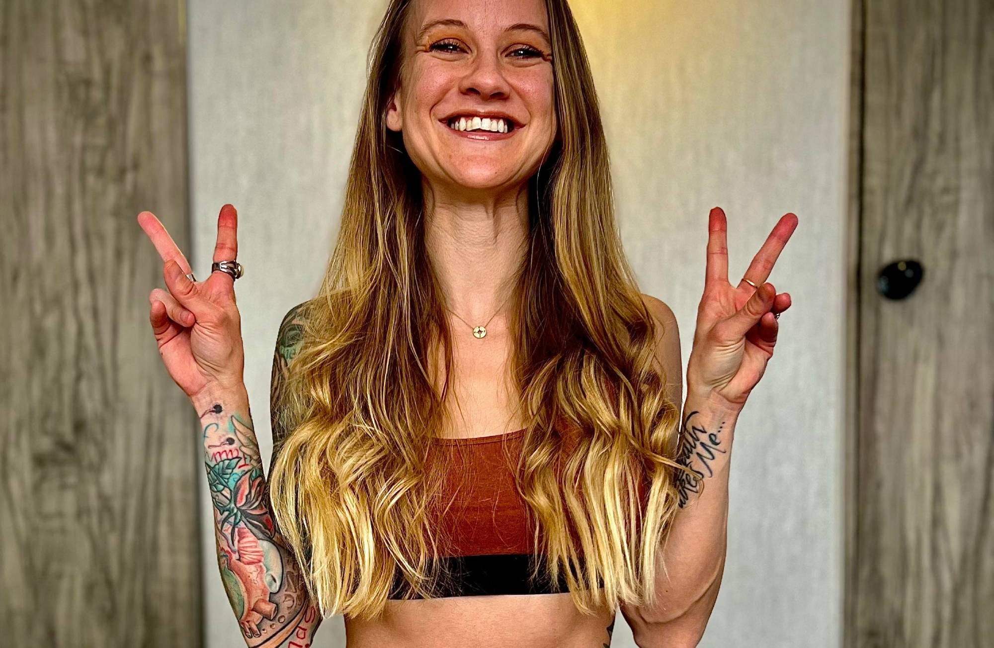  smiling blonde throws up two peace signs in a dark brown bralette