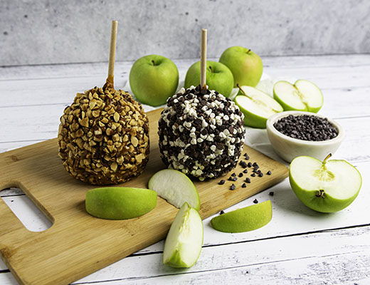 Two caramel apples on a cutting board with green apple slices.