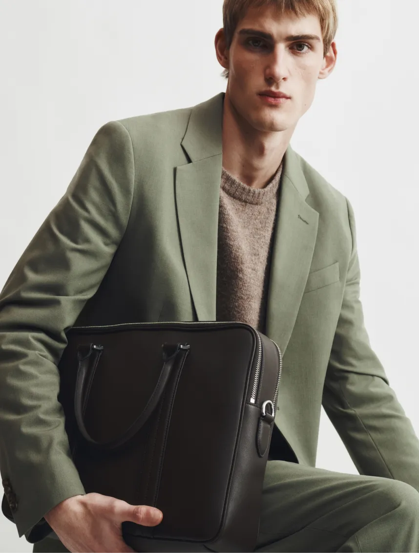 Young man in a green suit holding a black bag, sitting against a white background.