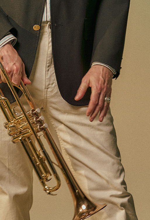 Person holding a trumpet, dressed in a blazer and light-colored pants.
