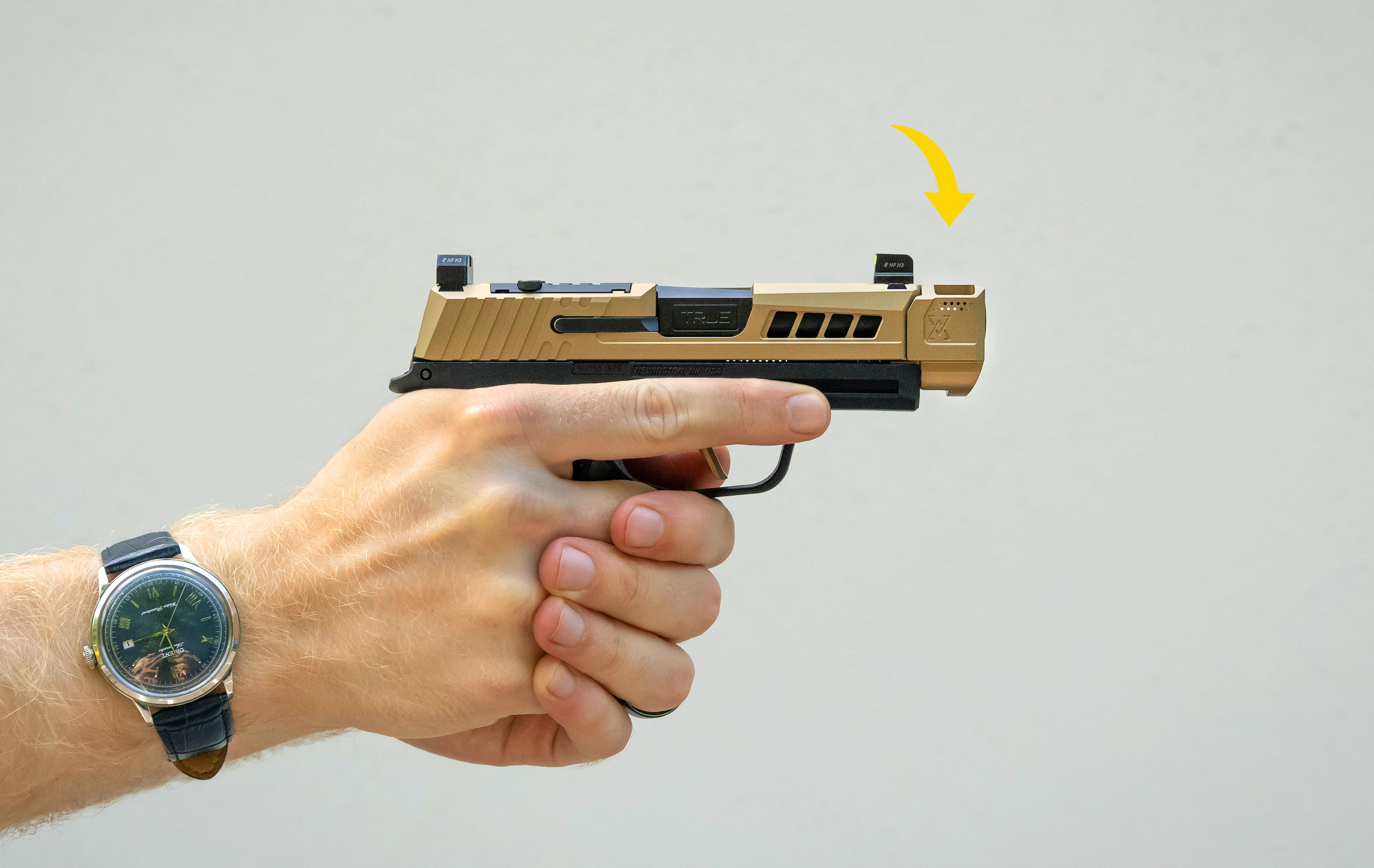 A hand holding a tan firearm with a watch on the wrist.