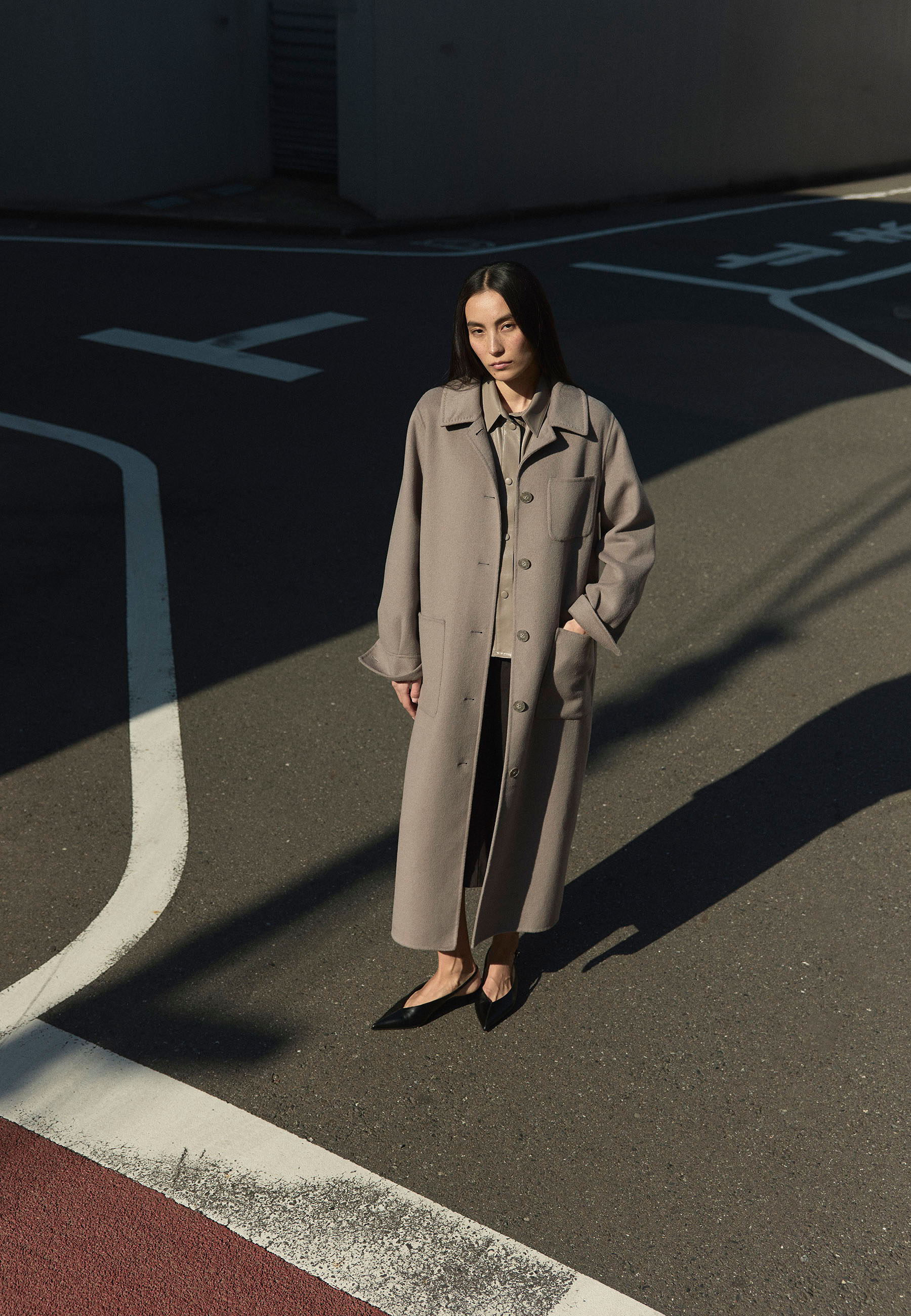 A woman in a gray coat stands on a street with shadows.