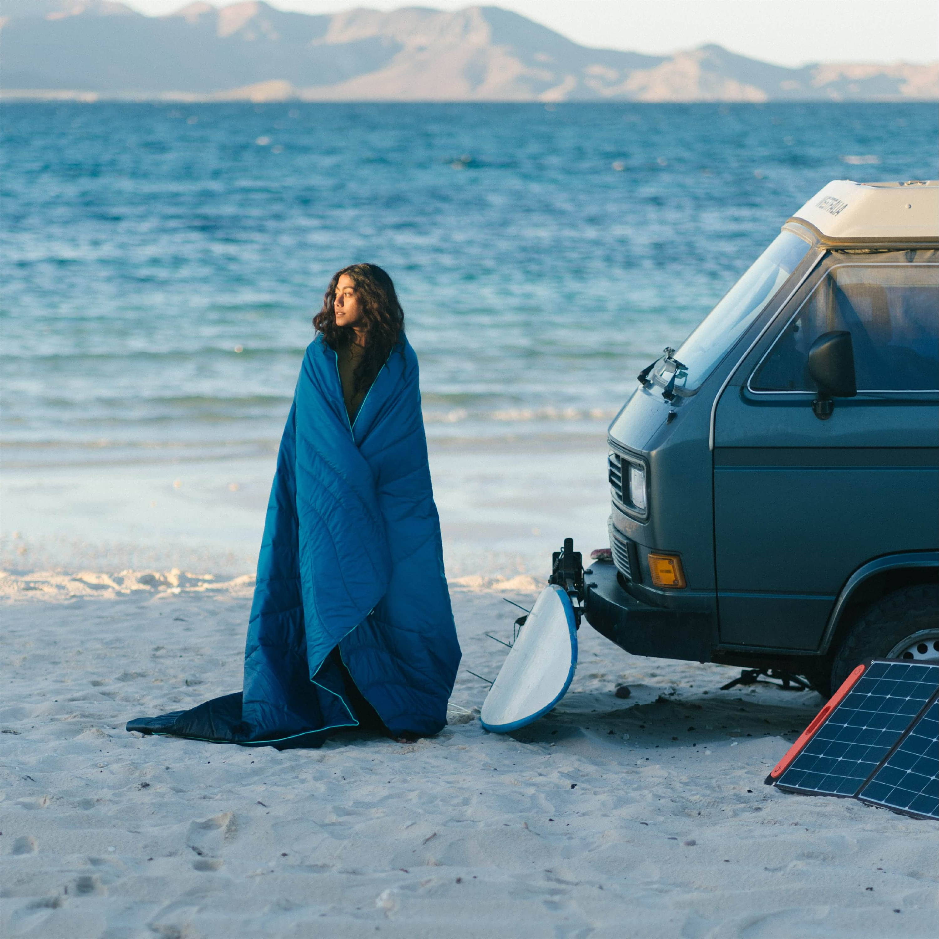 Woman Wrapped In Original Puffy Blanket On Beach In Front Of Camper Van
