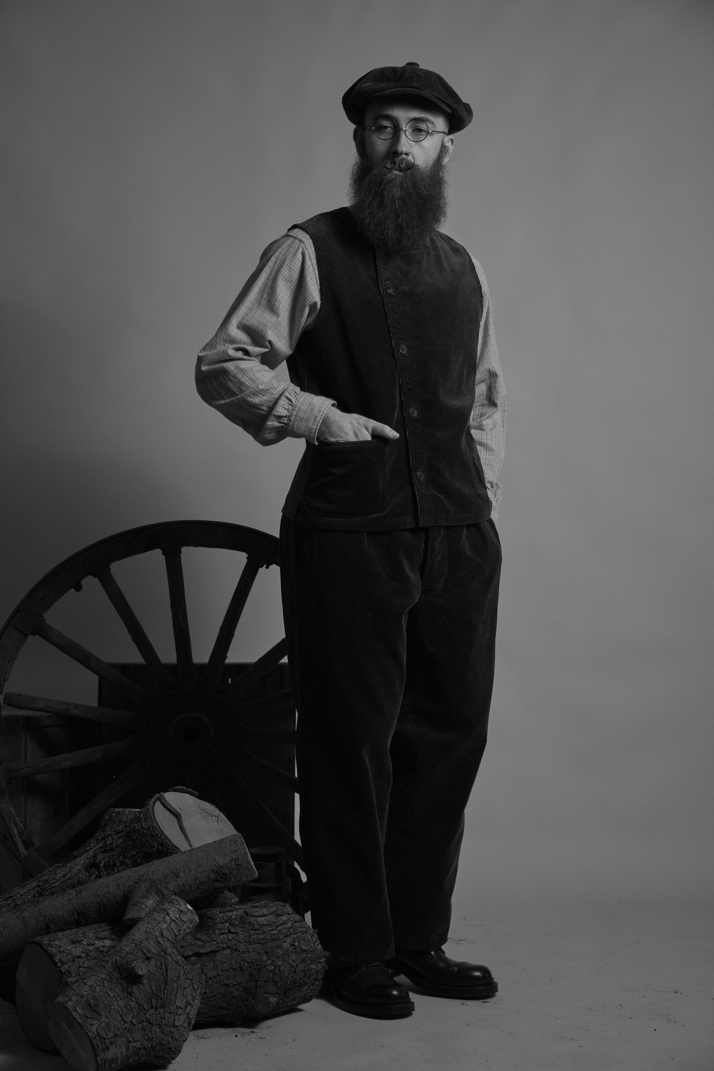 Man in vintage clothing standing near a wooden cart and logs.