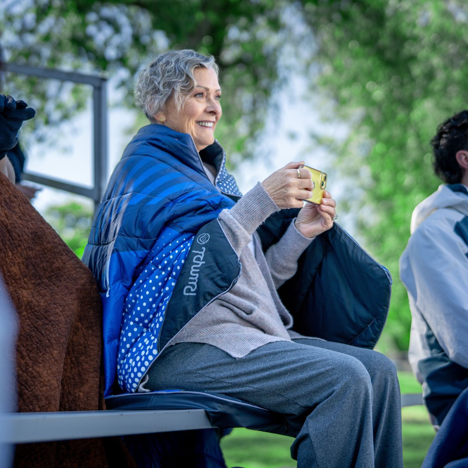 A woman sitting outdoors, wrapped in a blanket, smiling and holding a snack.