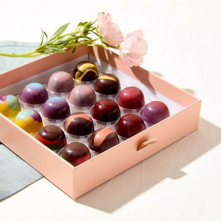A box of colorful chocolates with flowers beside it.