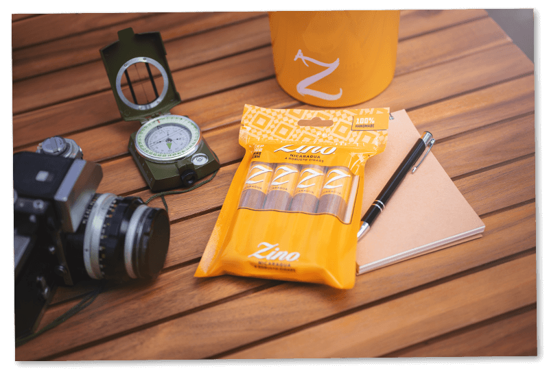 Four Zino Nicaragua Robusto cigars in their freshpack, placed on a wooden table alongside a note book, a pen, a Zino mug and a compass.