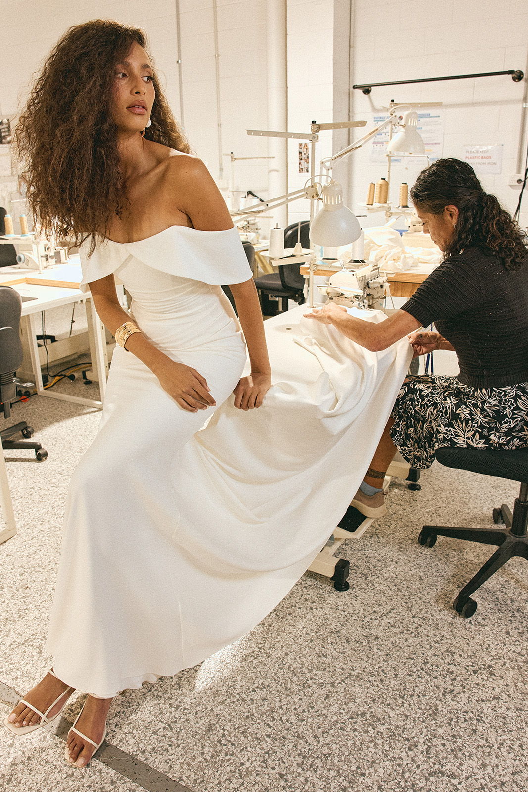 Model in a white dress fitting while a seamstress adjusts the hem.
