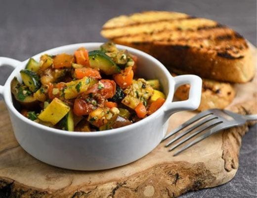 A bowl of vegetable stir-fry with grilled bread on a wooden board.