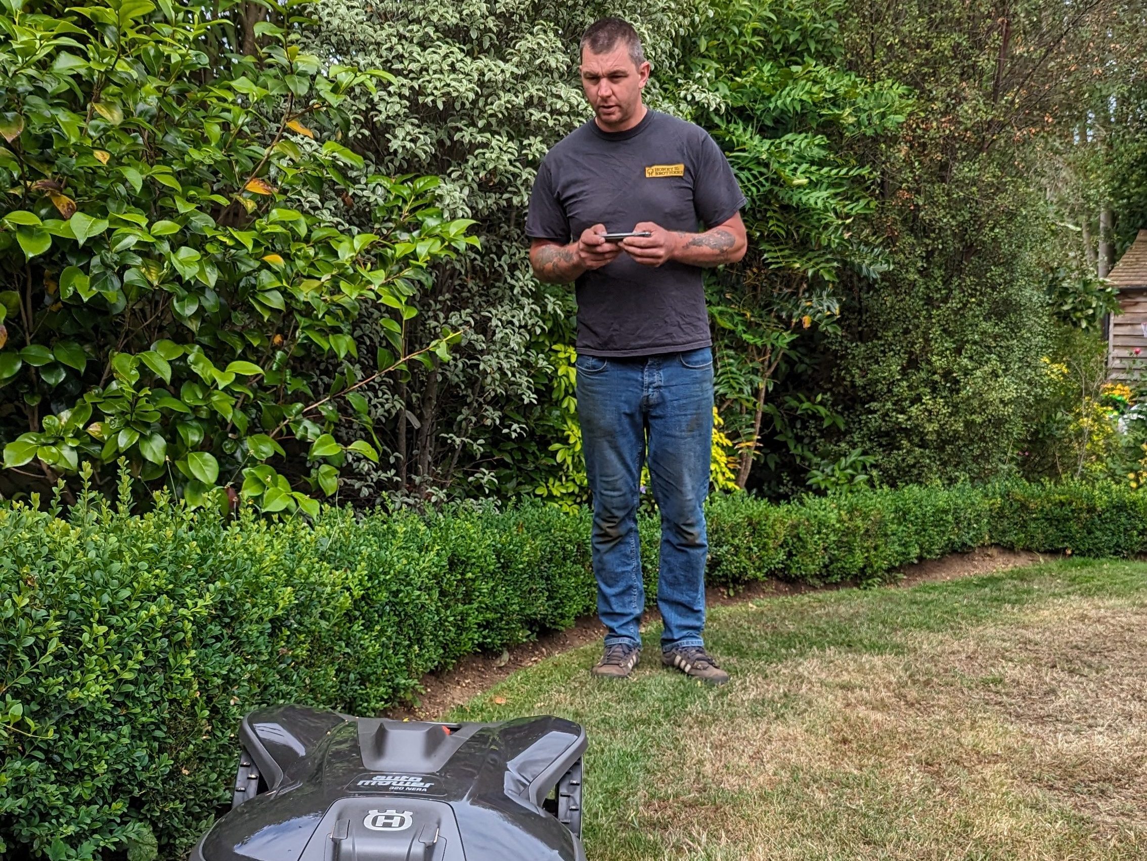 Man standing near a hedge, holding a device in a garden.