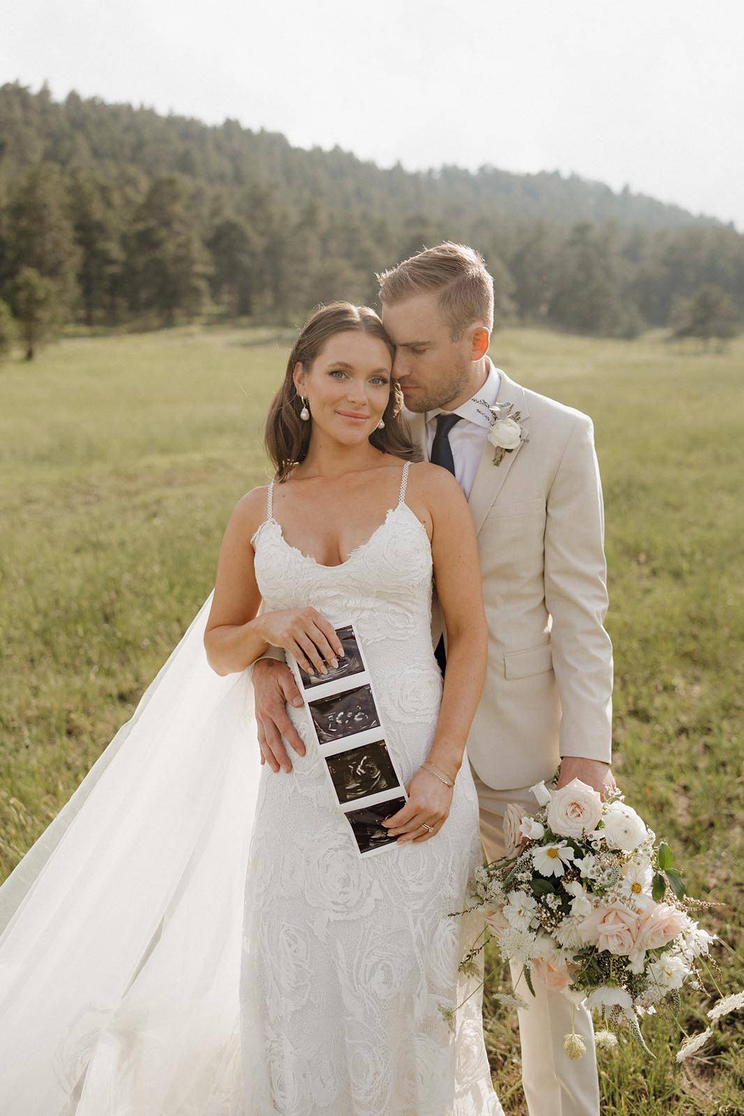 Pregnant bride, hand on belly, smiling and showing us a photograph of her baby bump