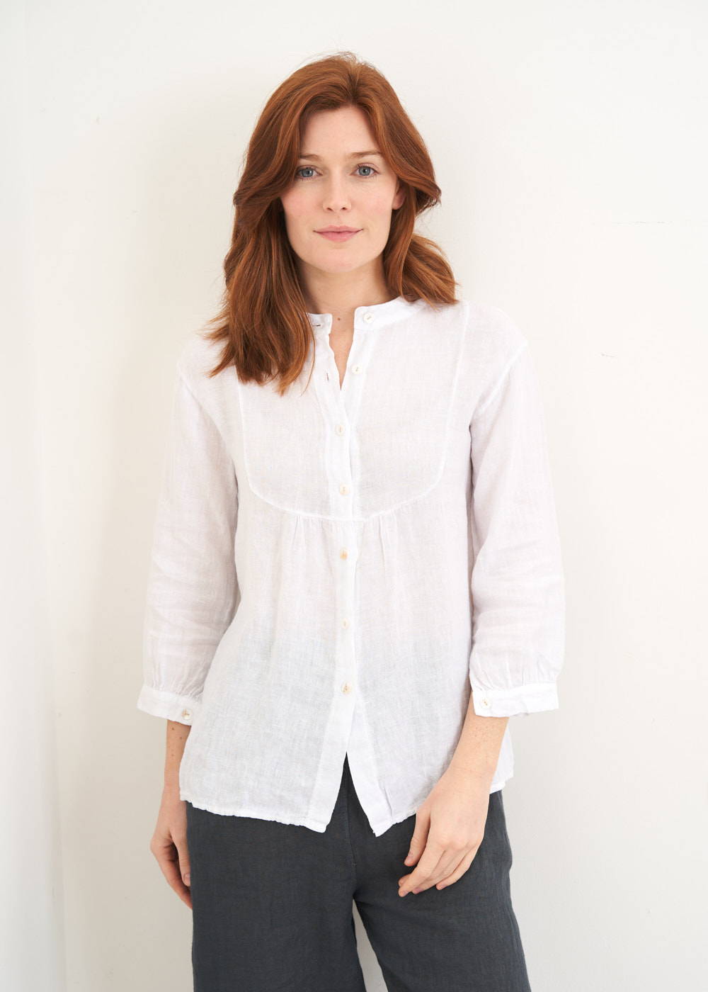 A model wearing a long sleeved white linen top with bib detail on the neckoine with black trousers and off white clogs