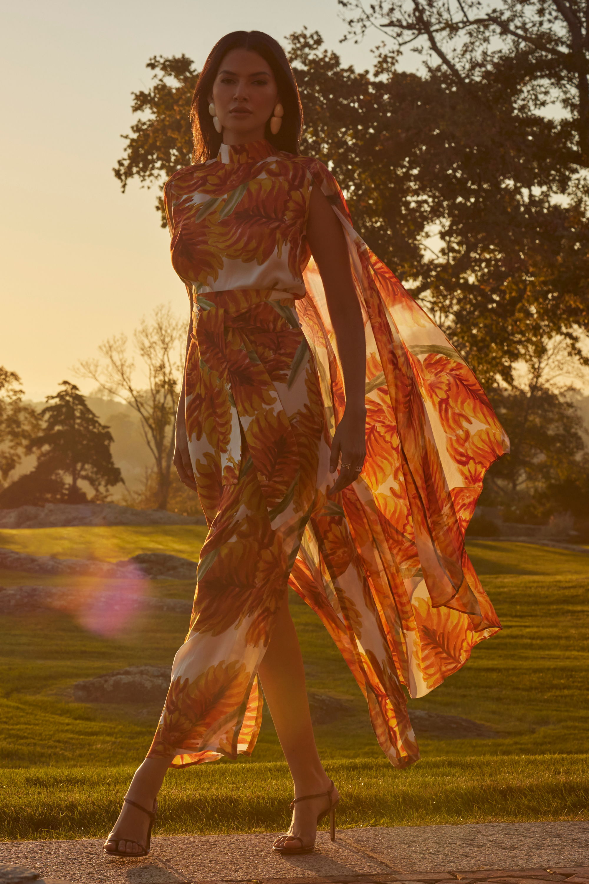 A woman in a flowing orange dress walks in a sunlit outdoor setting.