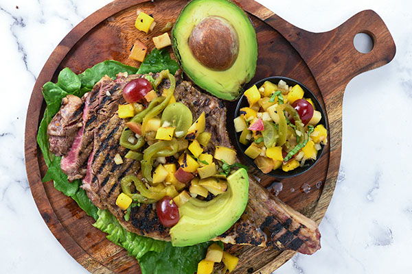 Grilled steak with avocado, peppers, and mango salsa on a wooden board.