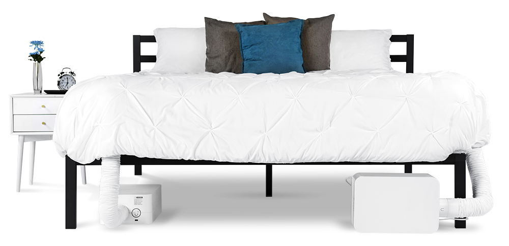 Bed with white bedspread and white, gray, and blue pillows, nightstand with flowers, alarm clock and remote control, and two BedJet units underneath the bedframe.
