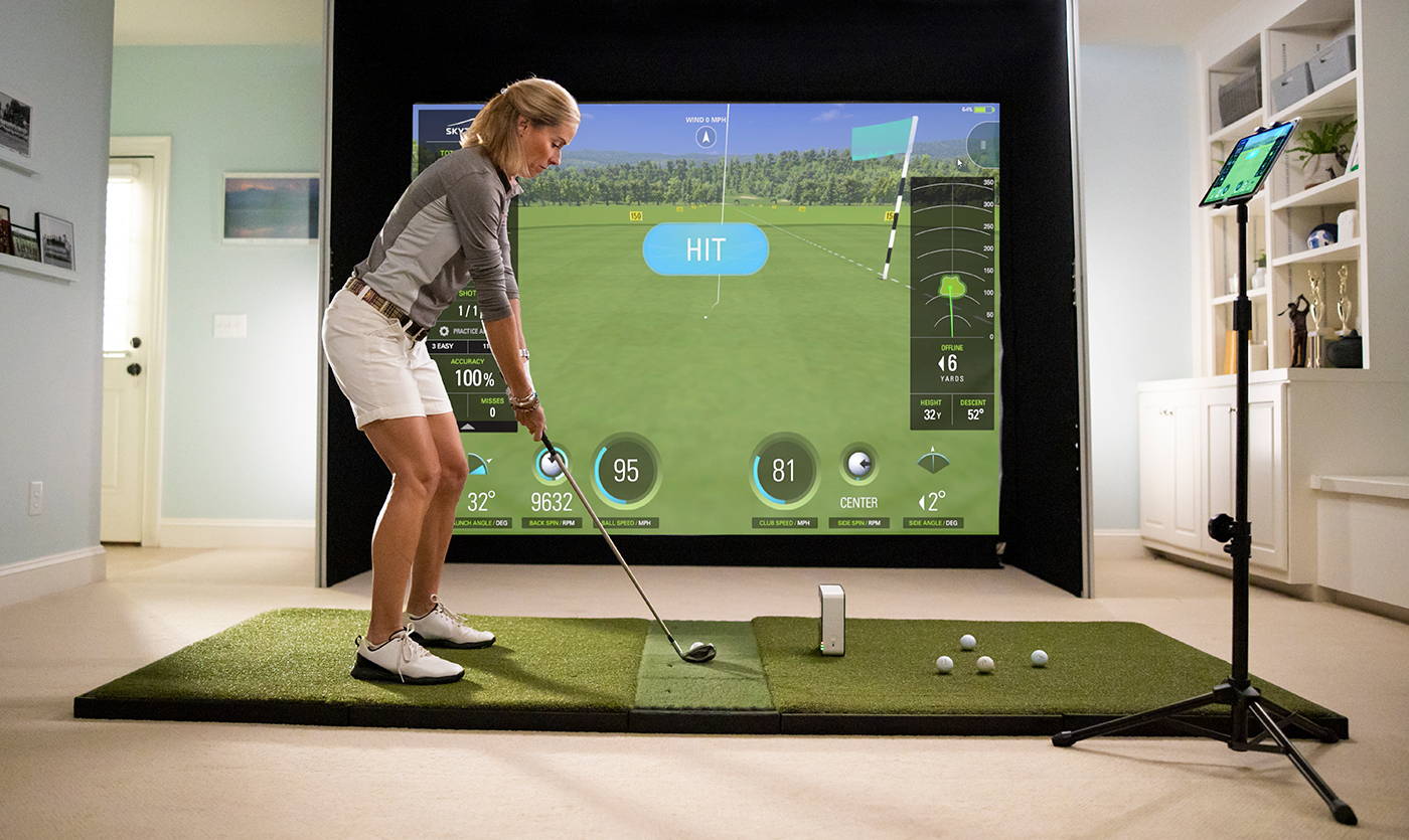 A woman addressing the golf ball next to a SkyTrak unit in a home golf simulator with a golf mat, impact screen and iPad on a stand