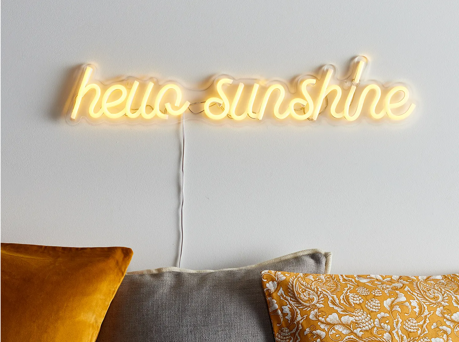 Neon 'hello sunshine' wall sign above some yellow and grey cushions.