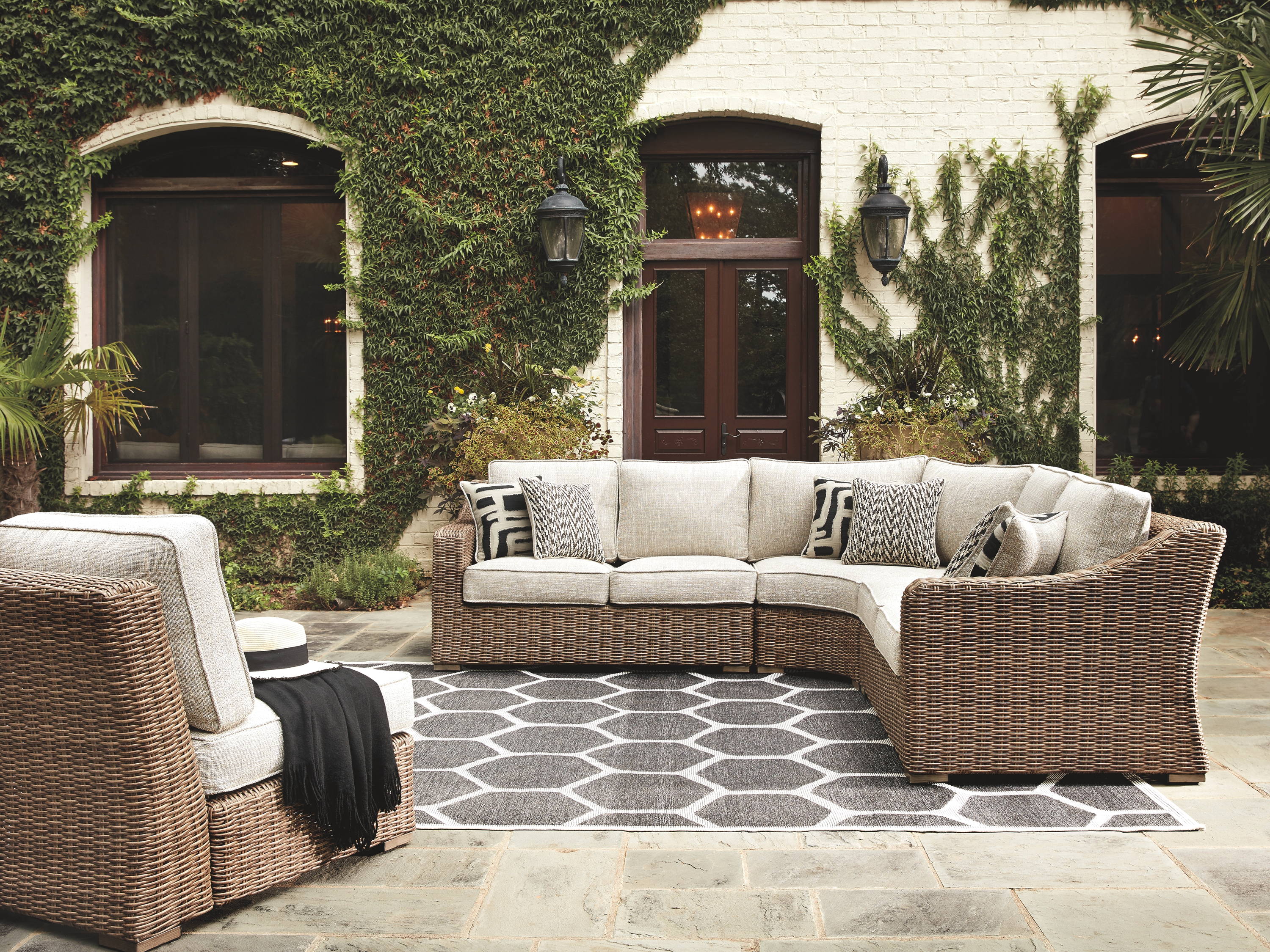 Outdoor sectional and chair sits on a hexagon rug with  a  greenery-filled background.