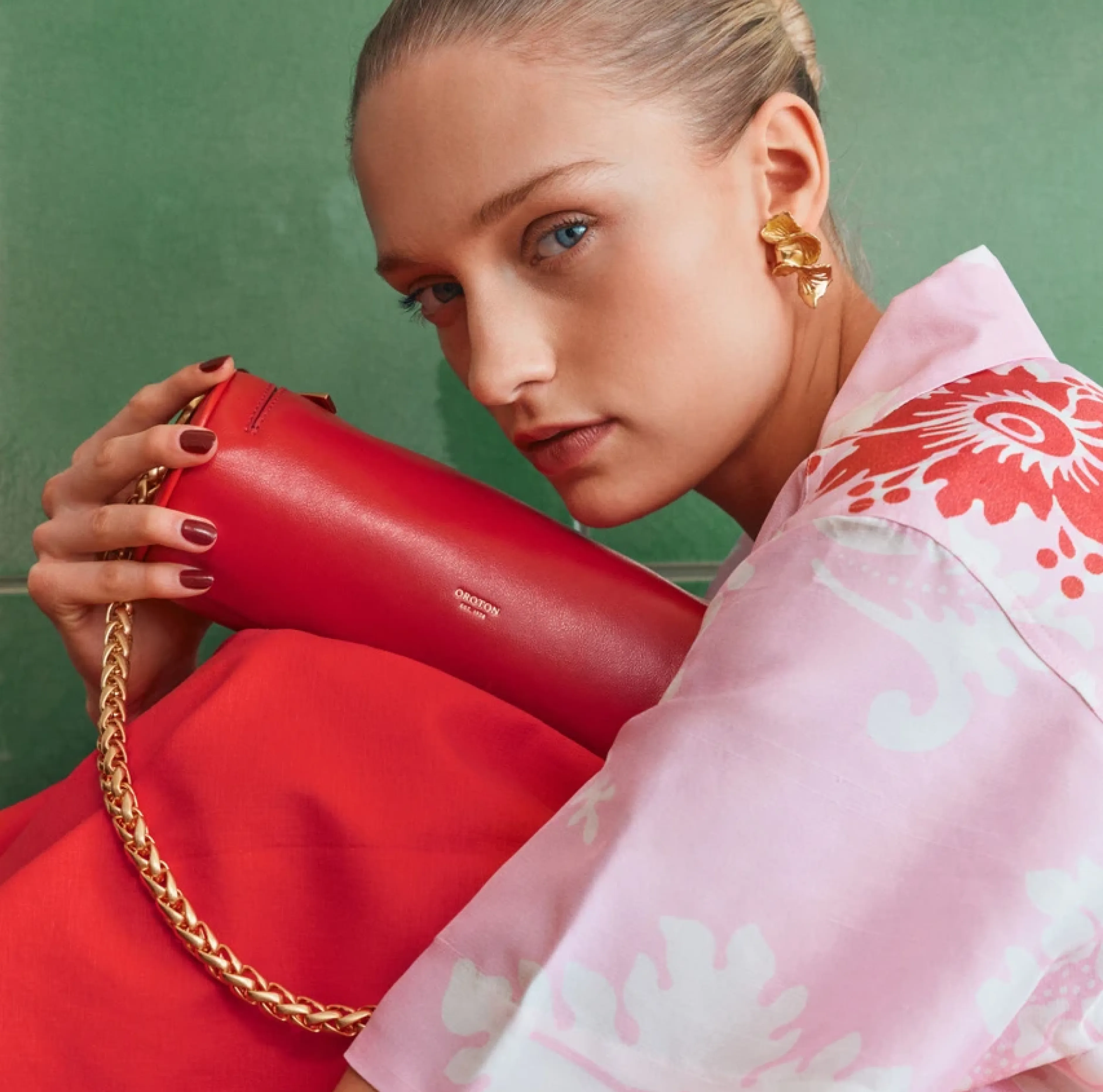 Woman holding a red handbag, wearing a floral shirt against a green background.
