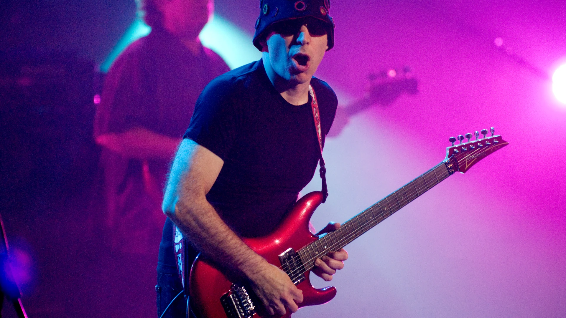 Joe Satriani playing electric guitar on stage with colourful lighting.