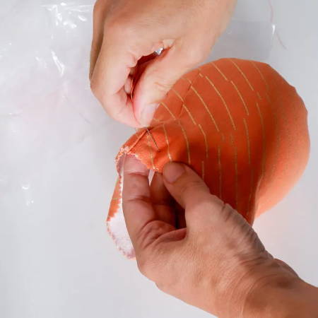 Hand sewing stitches around the opening of a stuffed orange fabric bowl-shaped piece