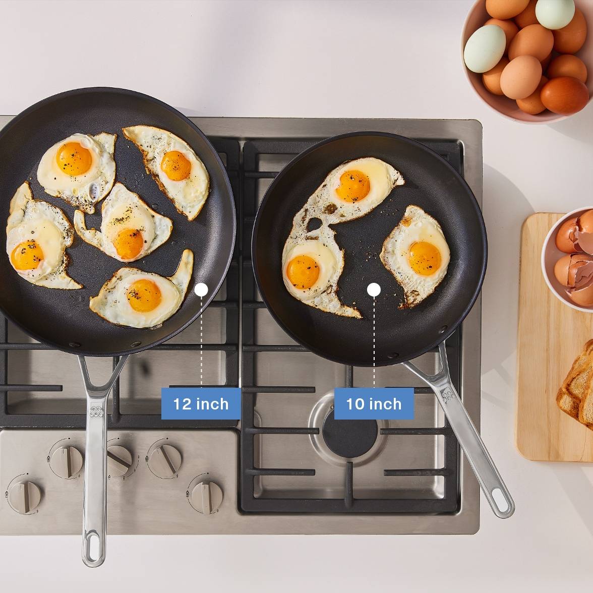 A bird’s eye view of the Misen 12-inch and 10-inch Nonstick Pans on a stovetop with fried eggs inside. To the right is a Misen Wood Cutting Board with toast and empty eggshells on it. A caption shows the 12-inch pan on the left, and the 10-inch pan on the right.