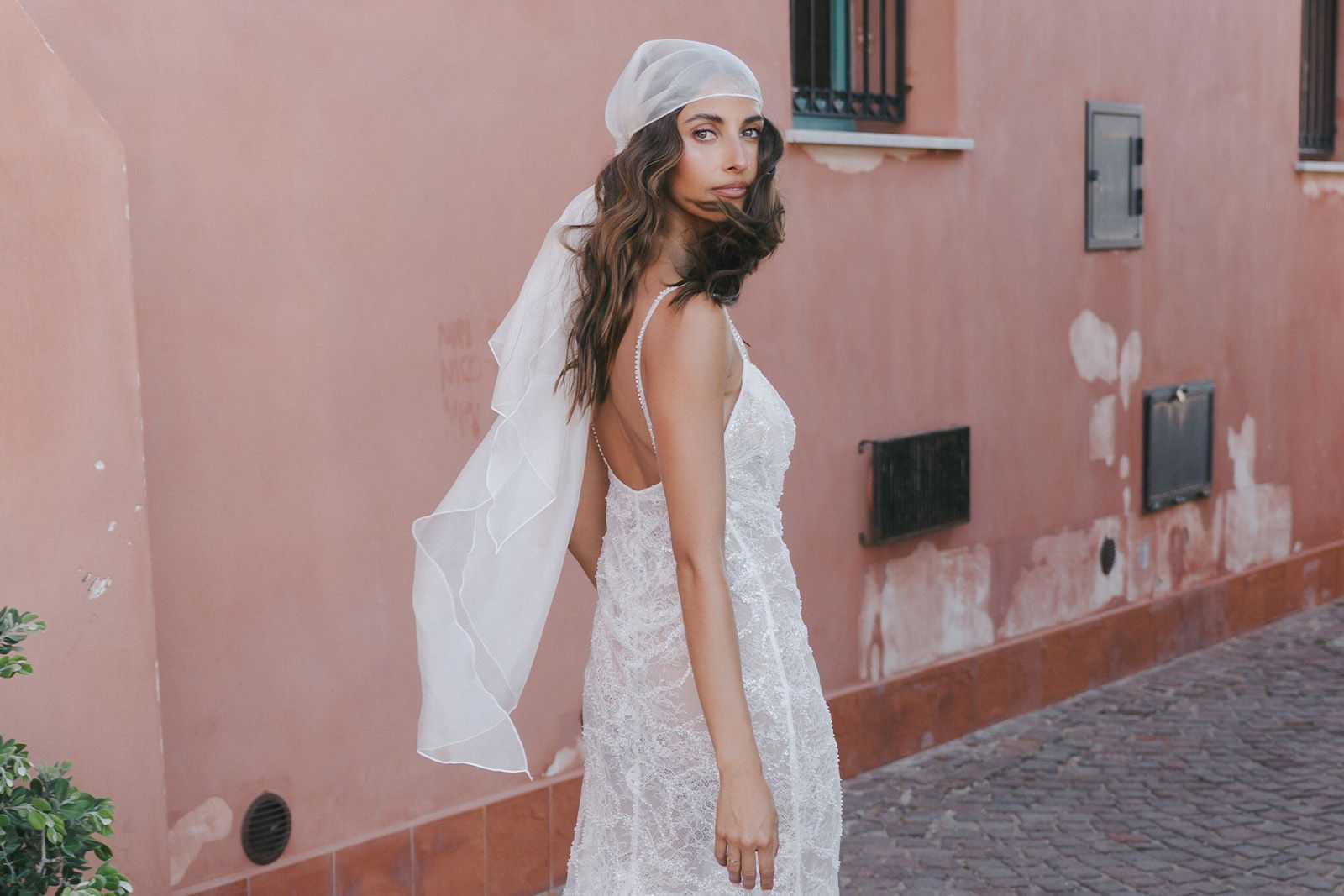 Woman in a white dress and headscarf standing against a pink wall.