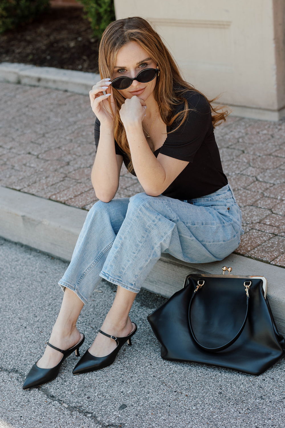 Young woman sitting on pavement, wearing sunglasses and casual outfit, with handbag.