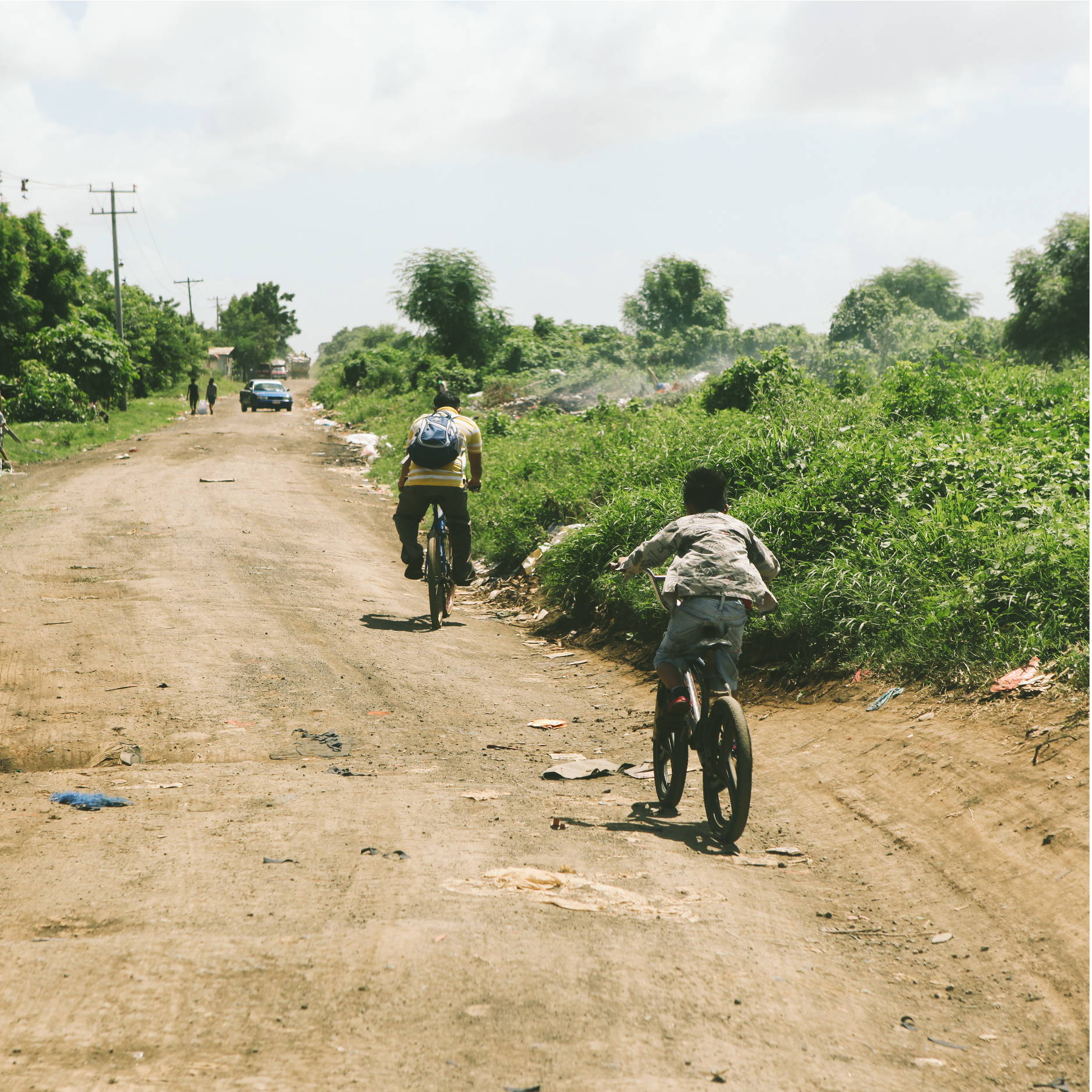 A man and his son ride their bikes down a trash lined dirt road in Nicaragua. The road is surrounded by overgrown vegetation.