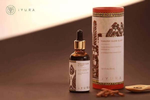 Una hermosa foto de Manjish Glow Elixir, el aceite de noche más vendido de iYURA, acompañado de su empaque cilíndrico protector bellamente ilustrado.