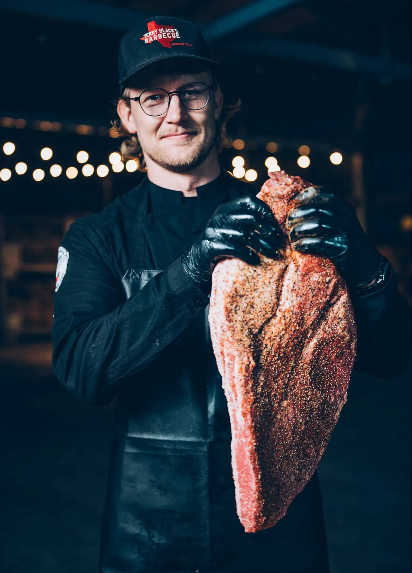 Person holding a large piece of seasoned meat, wearing glasses and a cap.