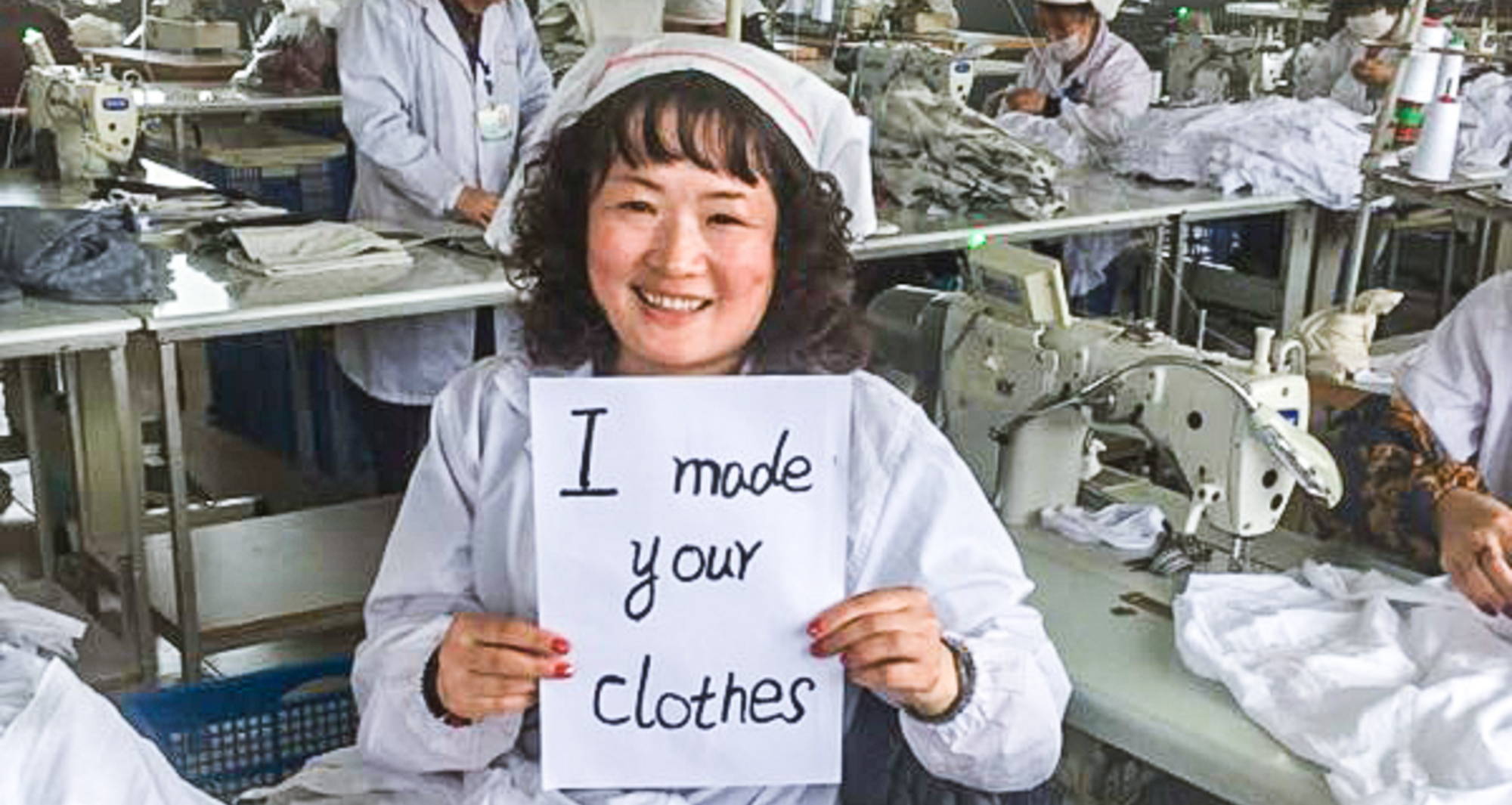 a woman in all white holds a sign that says “I made your clothes” in a clothing factory