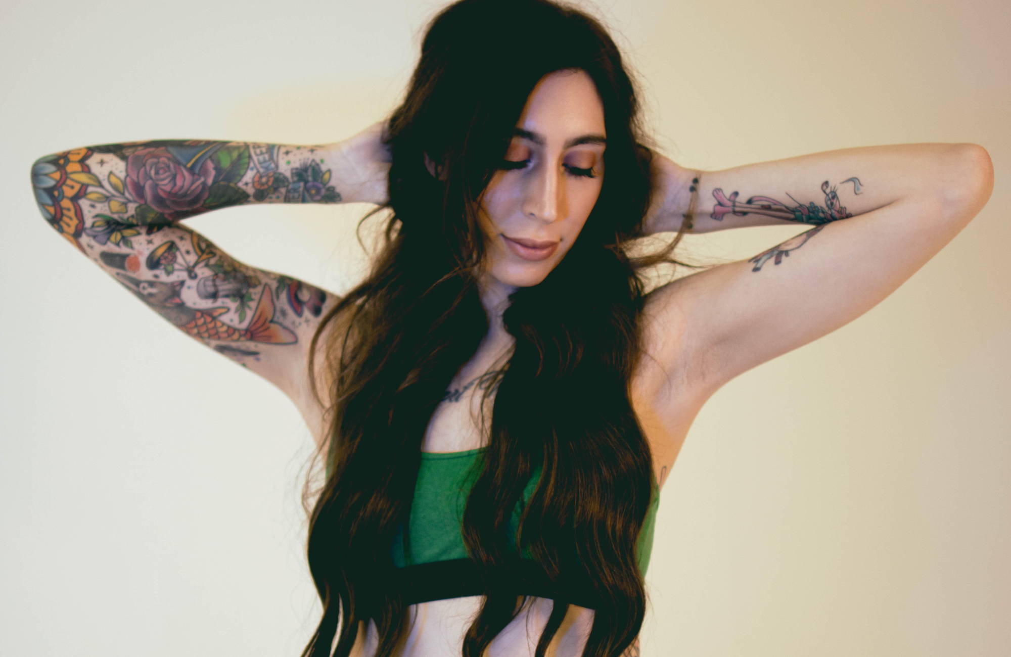 woman with long, dark hair has her hands on her head and looks down while wearing a green bralette